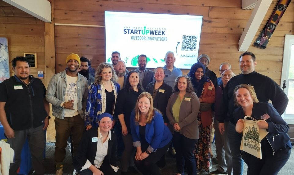 A group of people posing indoors with a screen displaying "Northern Michigan Startup Week: Outdoor Innovations" in the background.